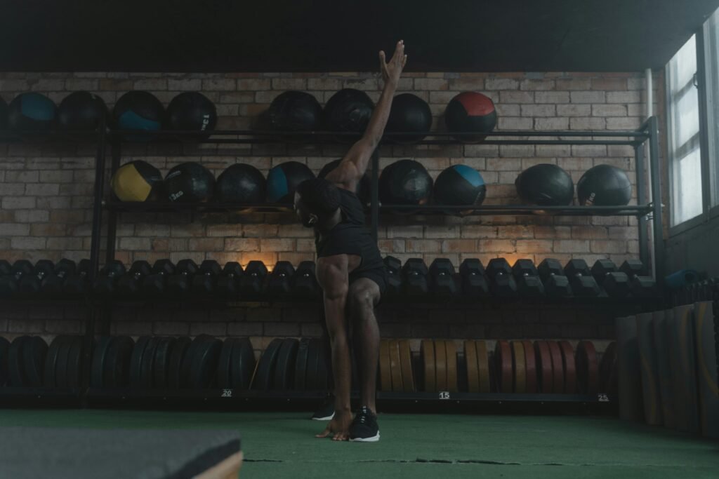 man stretching in a gym with medicine balls, dumbbells and plates behind him